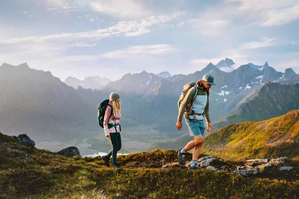 Zwei Wanderer mit Rucksäcken wandern bei Sonnenaufgang entlang eines grasbewachsenen Bergkamms, im Hintergrund sind schroffe Gipfel und ein bewölkter Himmel zu sehen.