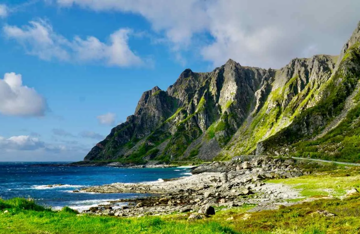 Spettacolare cartolina della costa norvegese che mostra ripide montagne ricoperte di vegetazione verde e baciate dalla luce del sole in cima, con le onde del mare ai piedi delle montagne.