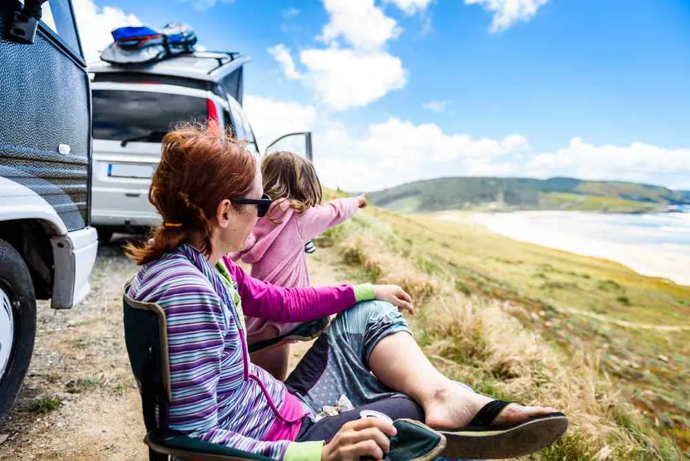 Zwei Frauen sitzen neben einem Van auf einer Klippe an der Küste; eine von ihnen zeigt auf den Strand und die Hügel unter einem strahlend blauen Himmel.