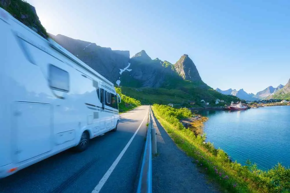 Ein weißer Campervan fährt auf einer sonnigen Küstenstraße in den Bergen entlang, vorbei an blauem Wasser und steilen grünen Gipfeln.