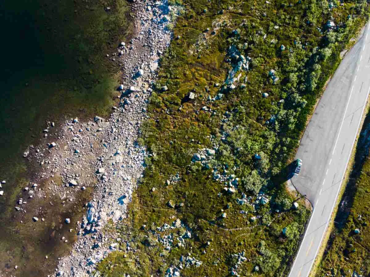 Luftfoto av en bil parkert langs riksvei 7 i Norge ved en steinete innsjøbredd og en grønn åsside.
