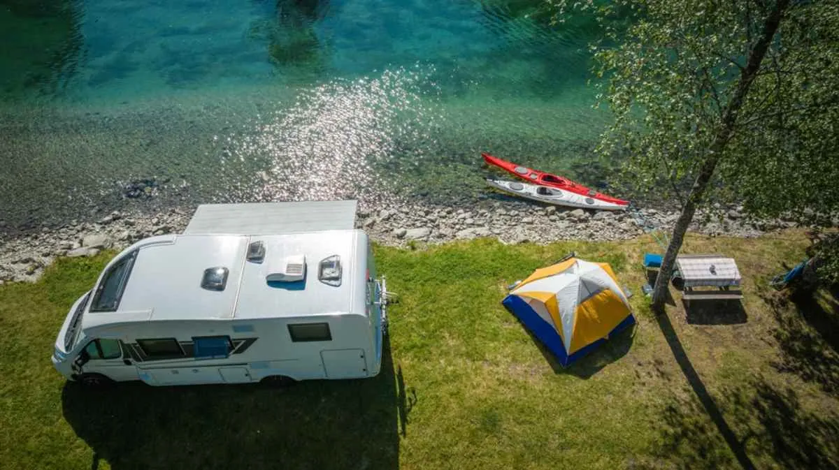 Camper parcheggiato in riva a un lago turchese con una tenda, un tavolo da picnic e un kayak, in un posto da campeggio panoramico e tranquillo immerso nella natura