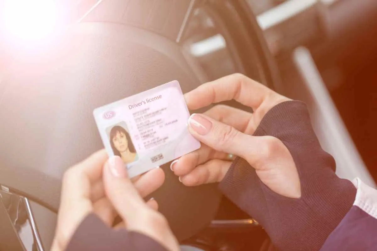 Una mujer con guantes sostiene un carné de conducir estadounidense mientras está al volante de un vehículo.