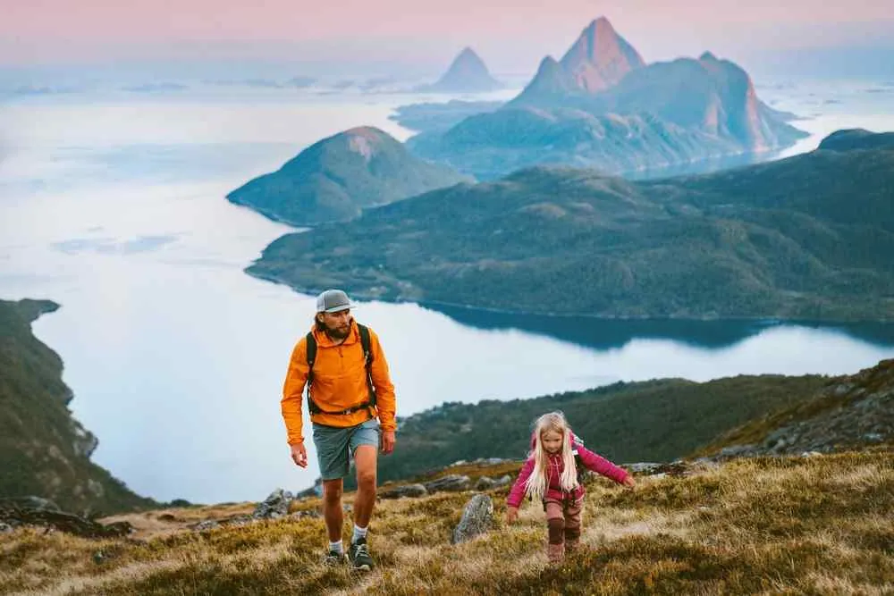 Ein Mann und ein junges Mädchen wandern einen grasbewachsenen Berg oberhalb eines Fjords hinauf, mit nebelverhangenen Gipfeln und ruhigem Wasser unter einem rosa-blauen Himmel.