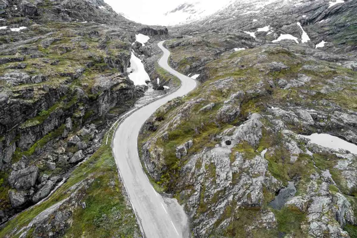 En svingete fjellvei i Norge som snor seg gjennom steinete terreng med snøflekker på begge sider.