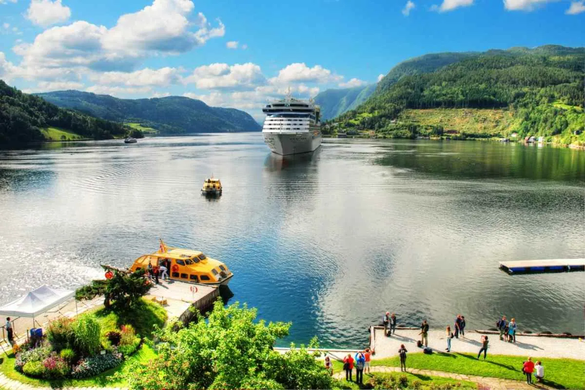 Kryssningsfartyg och tenderbåtar på Hardangerfjorden, sedda från en solig strandpromenad.