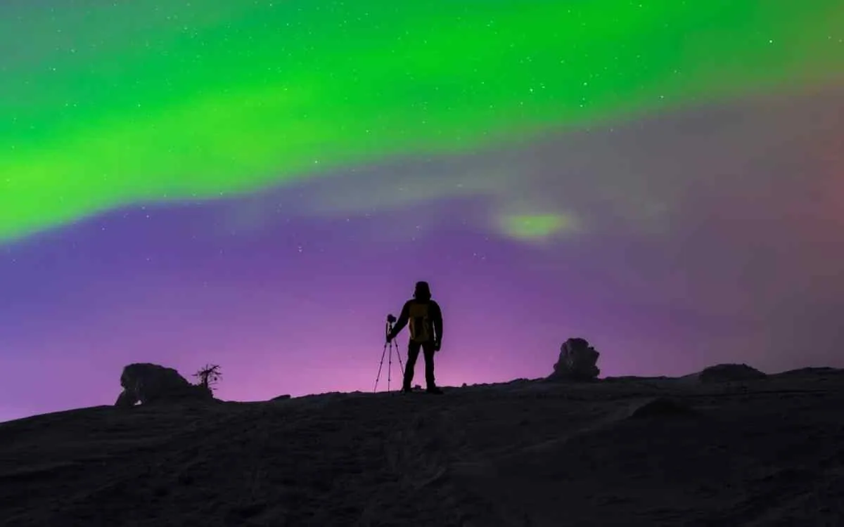 La sagoma di un fotografo su un crinale innevato vicino a Tromsø, mentre monta un treppiede sotto un’aurora verde brillante e un cielo notturno dai riflessi violacei.