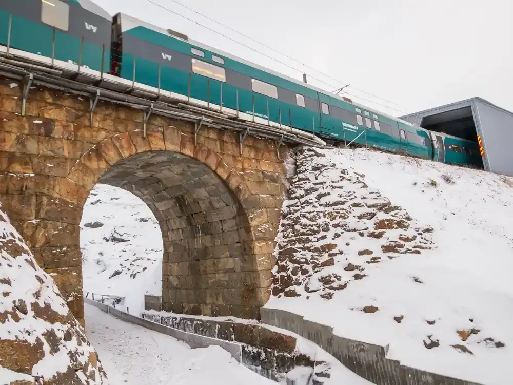 Et blågrønt tog krysser en steinbuebro over en snødekt kløft i et vinterlandskap.