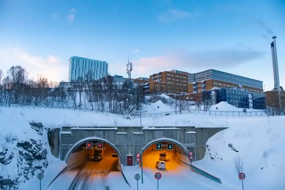 Snødekt bybilde med biler som kjører inn i to opplyste tunnelinnganger under leilighetsblokker og en skorstein i skumringen, 60 skilt synlige.