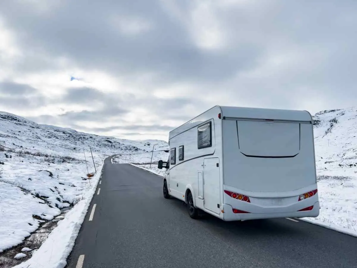 En vit husbil kör längs en asfalterad väg i Norge, omgiven av snö.
