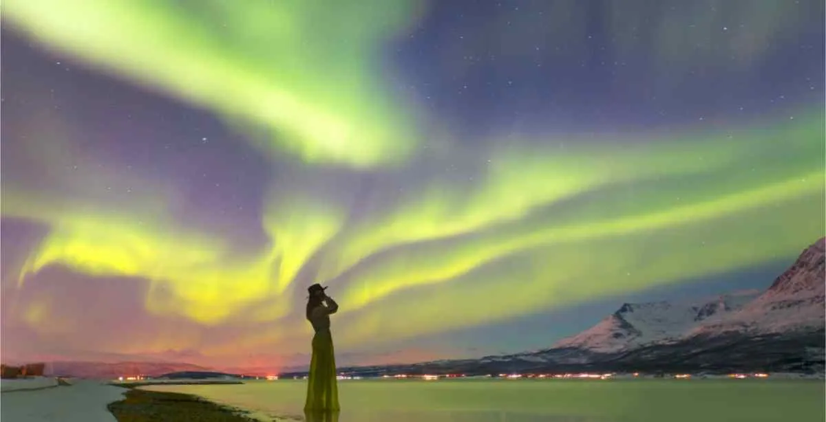 Una persona in riva a un fiordo vicino a Tromsø osserva l’aurora boreale giallo-verde che ondeggia sopra le montagne innevate e le luci lontane della città lungo la costa.