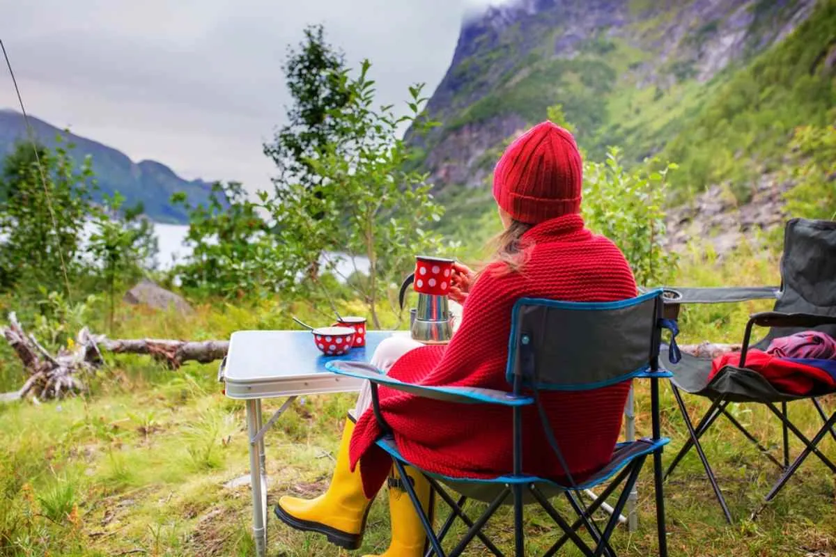 Personne vêtue d’un pull et d’un bonnet rouges, assise sur une chaise de camping et chaussée de bottes jaunes, en train de déguster un café au bord d’un fjord lors d’un séjour en camping sauvage en Norvège.