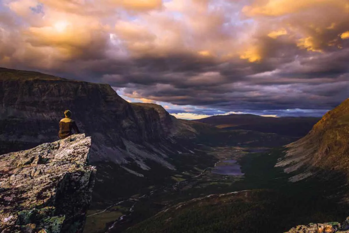 En reisende sitter på kanten av en klippe med utsikt over en dramatisk norsk dal ved solnedgang, med lagdelte fjell og en svingete elv nedenfor.