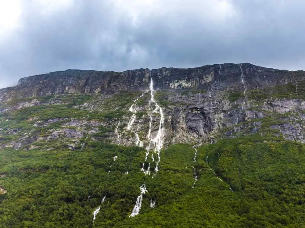 Zahlreiche Wasserfälle stürzen unter einem bewölkten Himmel eine steile Felswand hinab in einen üppig grünen Wald und schaffen so eine dramatische Bergkulisse