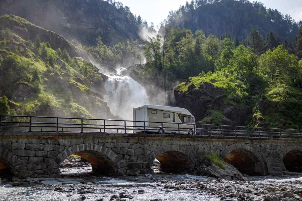 Un camper beige percorre una strada norvegese accanto a una cascata, passando su un ponte ad archi attraverso i quali scorre l’acqua.