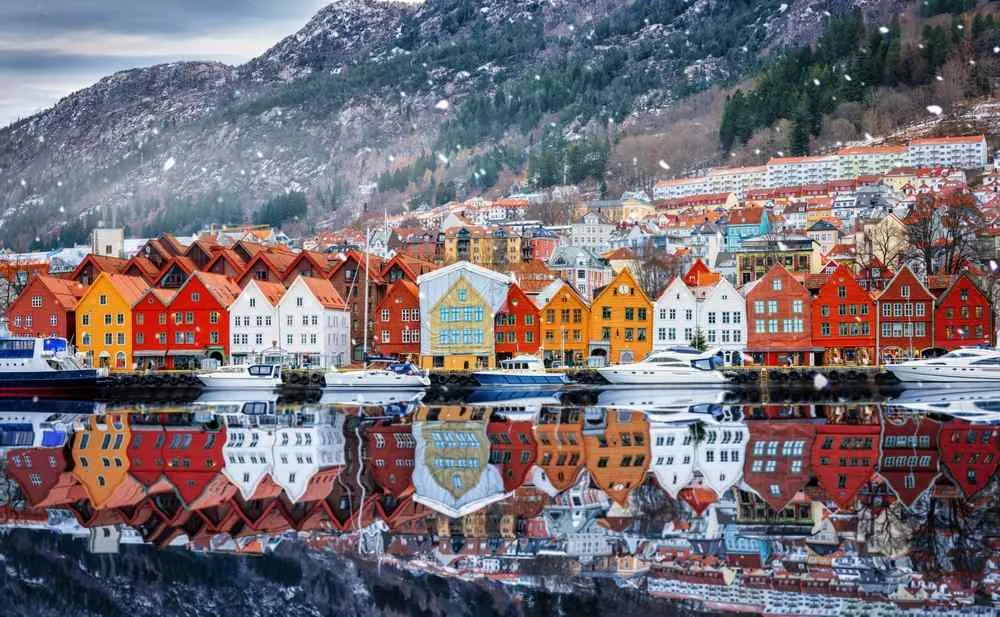 Farbenfrohe Häuser am Wasser und Boote in einer verschneiten Fjordstadt, in deren ruhigem Wasser sich die Berge spiegeln.