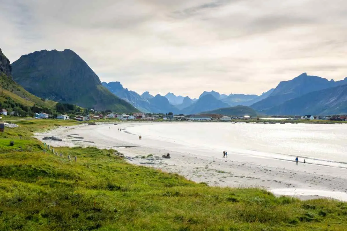 Rambergstranden på Lofoten, en vidstrakt bukt med hvit sandstrand hvor folk spaserer langs kysten, med spisse fjelltopper i bakgrunnen.
