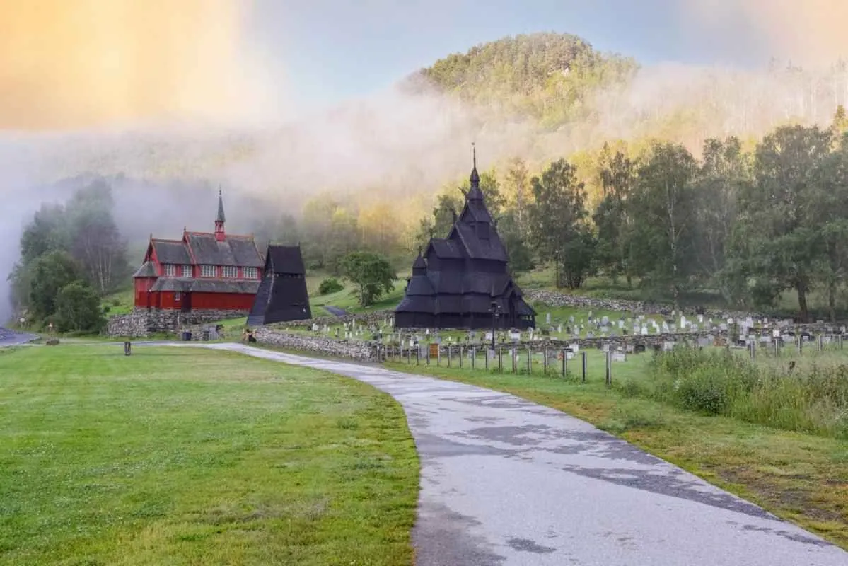 Die Stabkirche von Borgund in einem grünen Tal mit nebelverhangenen Hügeln, einem Friedhof und einem gepflasterten Weg, der zur Holzkirche führt.