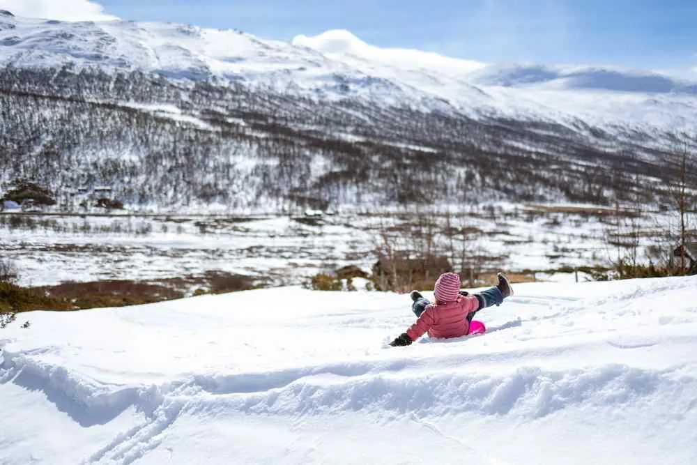 Ein Kind in einer rosa Jacke und einer gestreiften Mütze rutscht durch tiefen Schnee, im Hintergrund sind schneebedeckte Berge und ein blauer Himmel zu sehen.