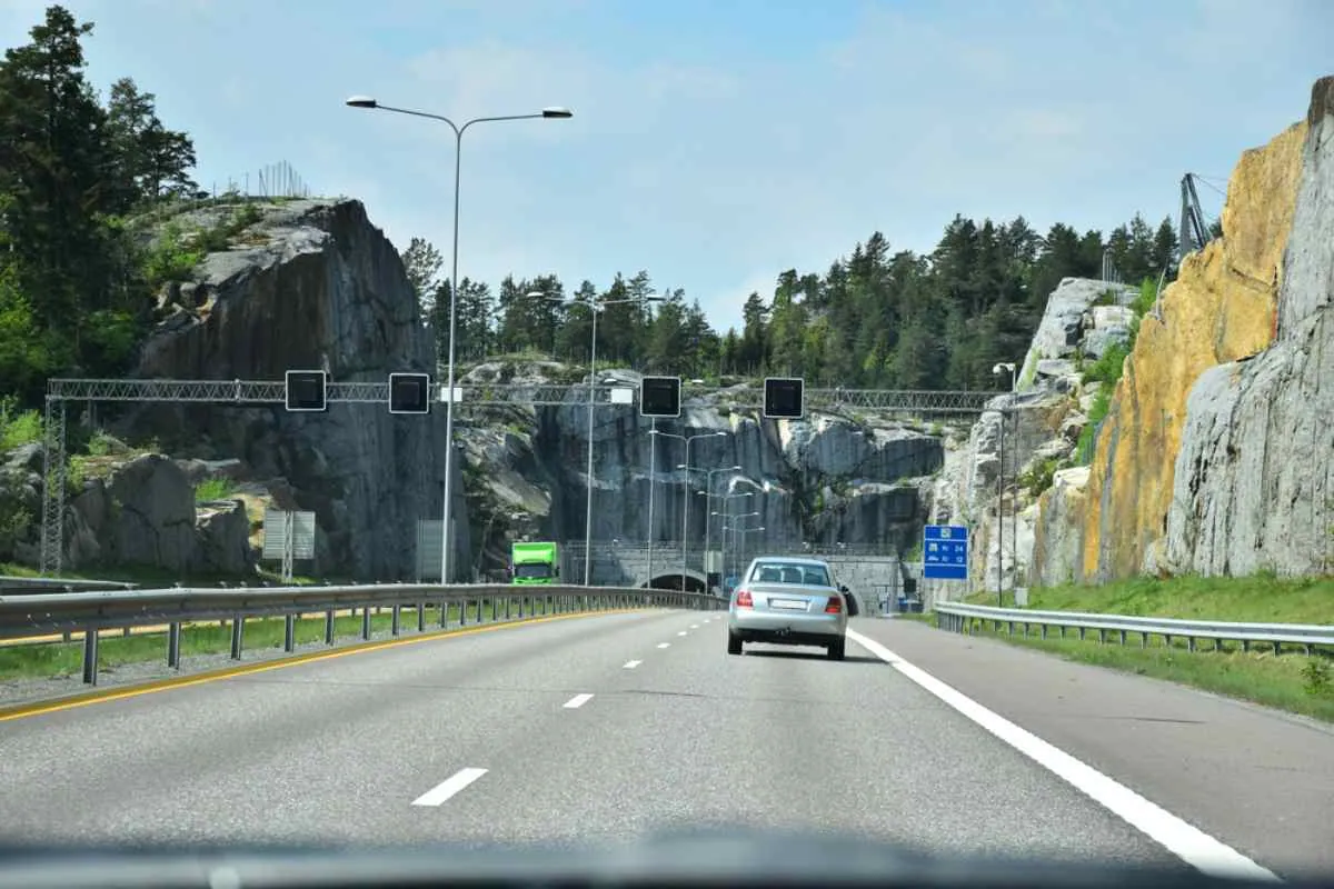 En firefelts norsk motorvei som fører inn i en steintunnel med elektroniske bomporter over veien – et typisk oppsett for automatisert veibompengeinnkreving.