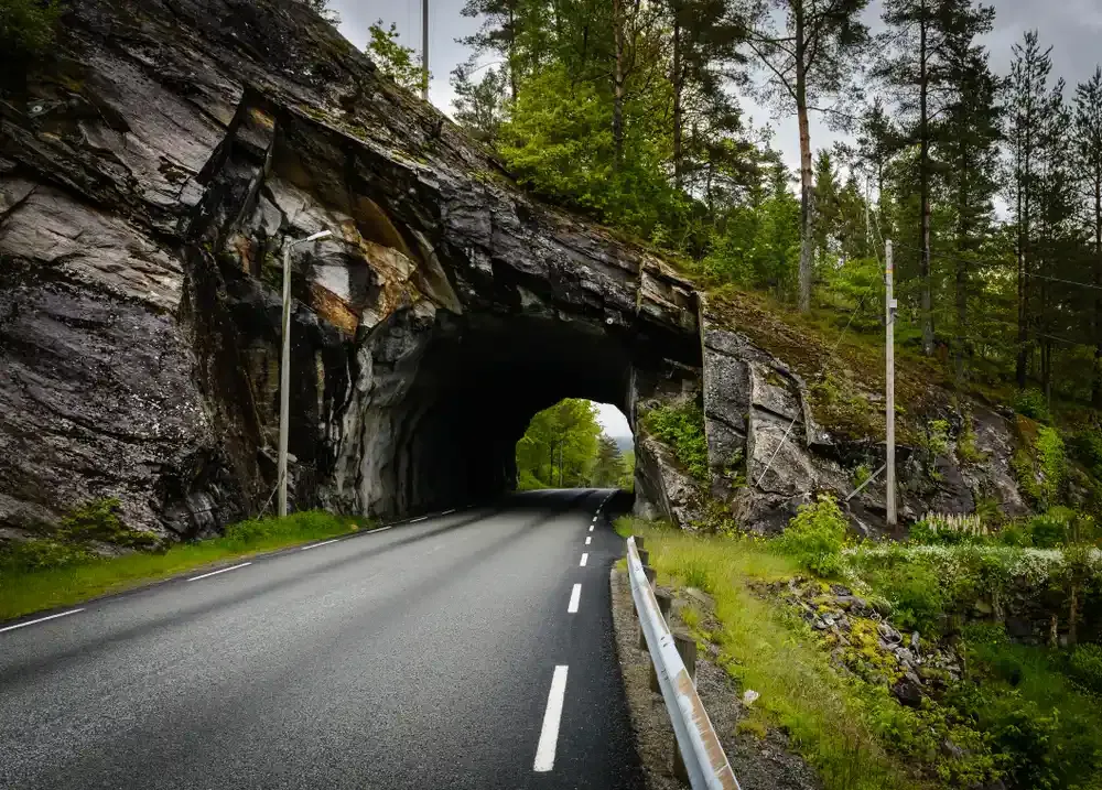 En tom vei som fører inn i en mørk tunnel hugget ut i en skogkledd klippe, med grønne trær, rekkverk og overskyet himmel.
