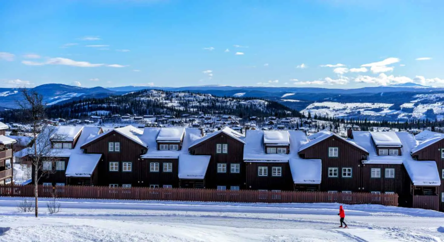 Schneebedeckte Hütten in Beitostølen mit einem einsamen Skifahrer auf einer Piste und blauen Bergen in der Ferne.
