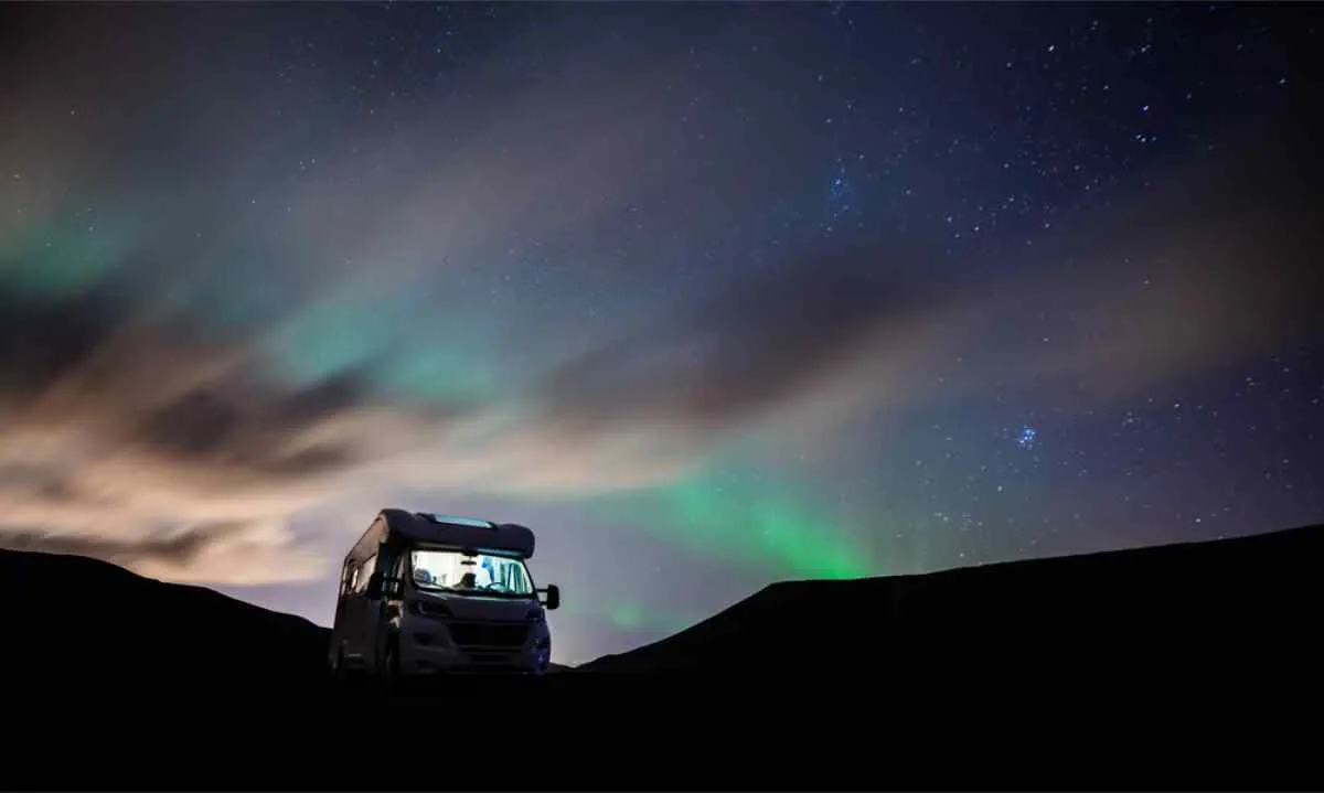 Camper parcheggiato sotto un cielo scuro e stellato vicino a Tromsø, con una tenue aurora verde che si estende sopra basse colline e nuvole fluttuanti.