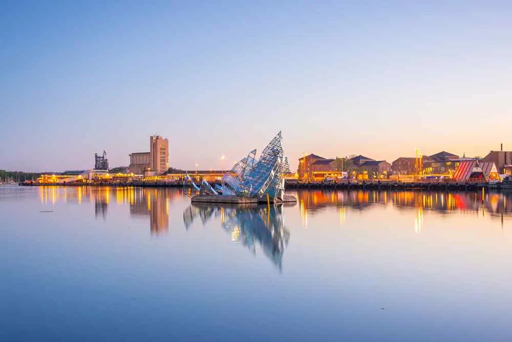 Die Skyline des Hafens in der Abenddämmerung mit einer Glasskulptur auf ruhigem Wasser, während sich die Lichter der Stadt im friedlichen pastellfarbenen Himmel spiegeln