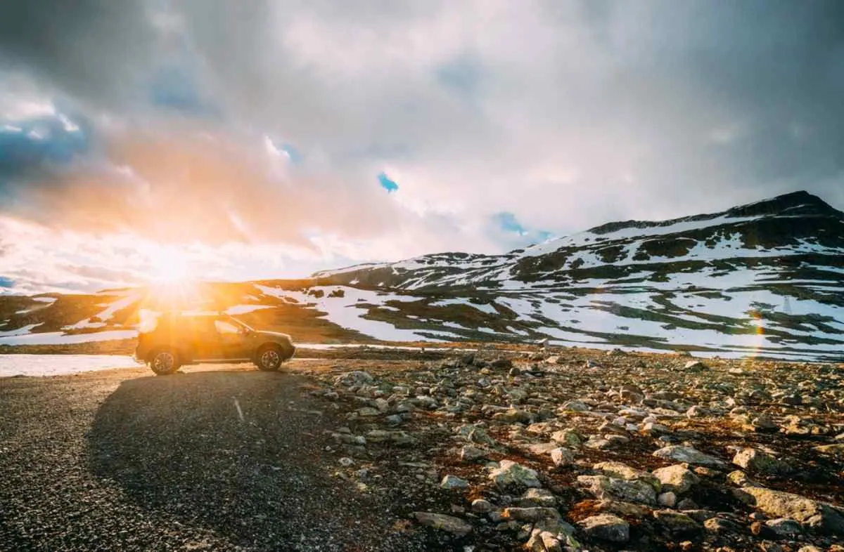 Un veicolo, illuminato dagli ultimi raggi di sole della giornata, rimane parcheggiato su una strada norvegese vicino a montagne innevate e grandi massi.