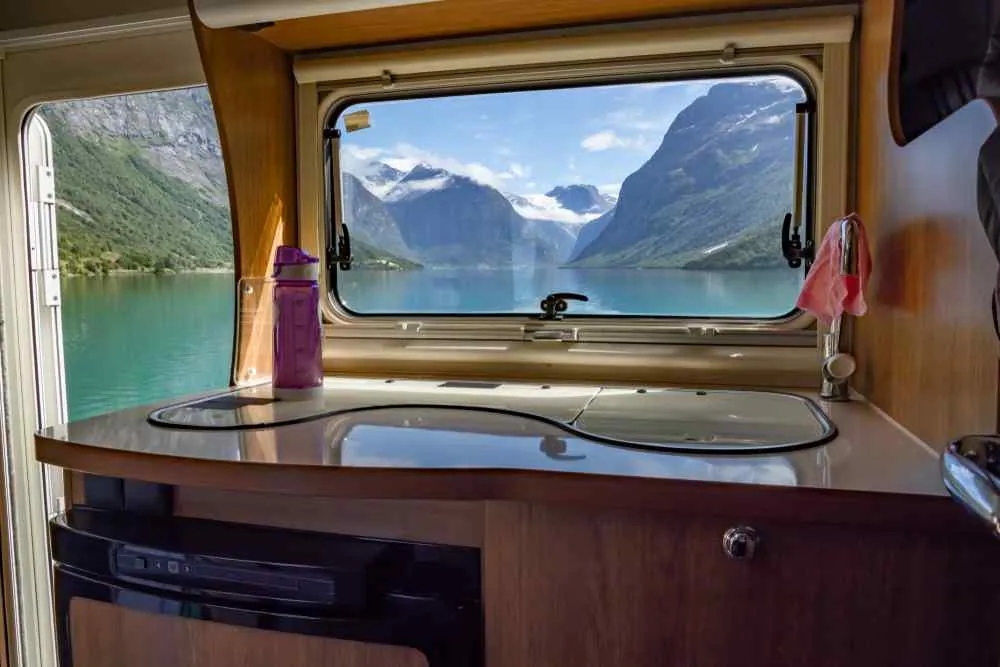 Küchenzeile im Wohnmobil mit einer violetten Flasche, Blick durch das Fenster auf einen türkisfarbenen Fjord und schneebedeckte Berge, ruhige, malerische Aussicht