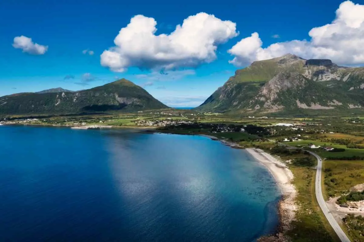 Vista dal drone di una parte della costa norvegese che mostra una strada vicino al mare e, sullo sfondo, due grandi catene montuose che sfiorano le nuvole bianche in una giornata dal cielo bluastro.