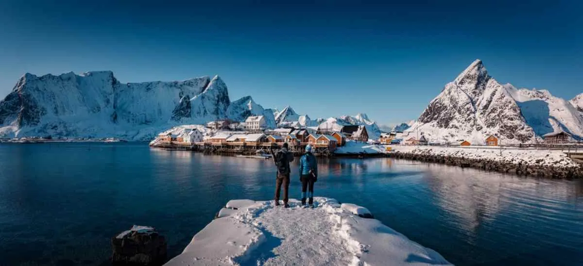Översiktsbild av en helt snötäckt bergskedja vid havet och en kuststad i norra Norge, medan en mur med många människor tornar upp sig på den andra sidan av vattnet.
