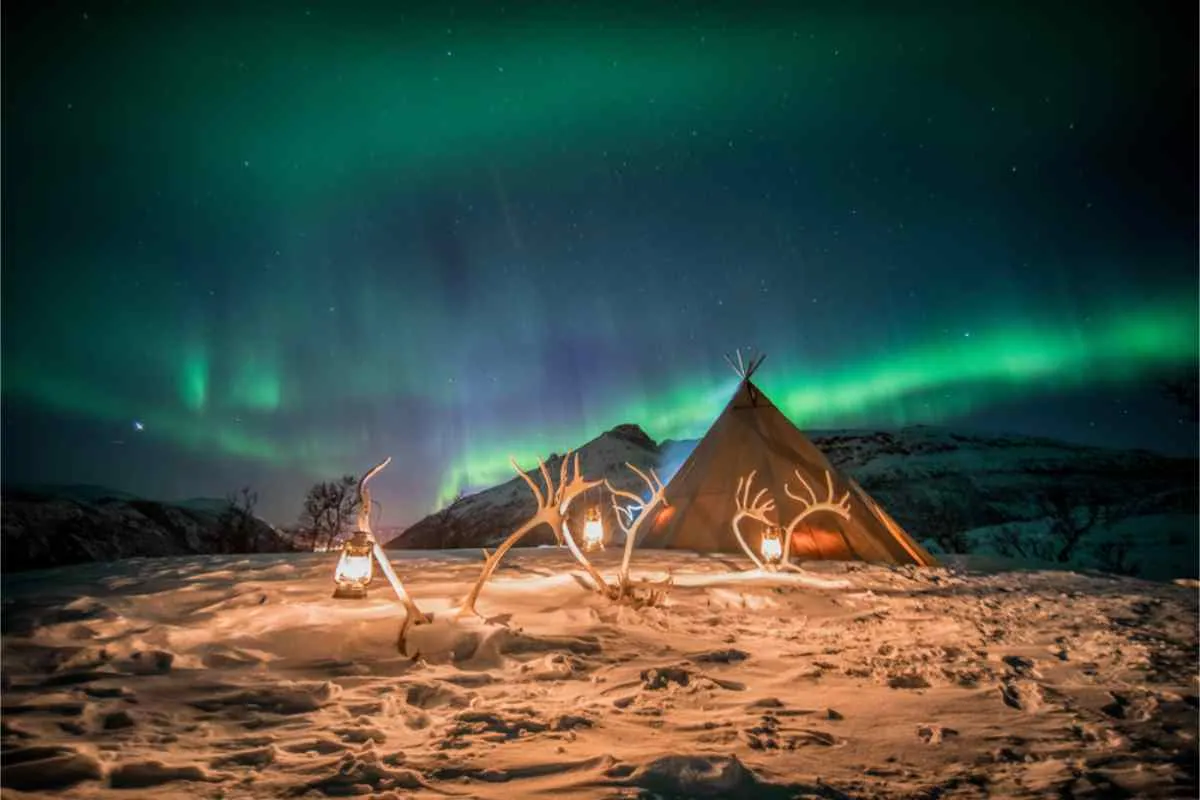 Un'aurora boreale verde sopra le luci della città e il lungomare di Tromsø, con la Cattedrale Artica che risplende in lontananza sotto un cielo stellato.