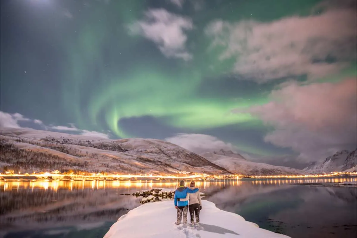 To personer står på snøen og betrakter nordlyset over en rolig innsjø og snødekte fjell, under en himmel i grønne og rosa nyanser.