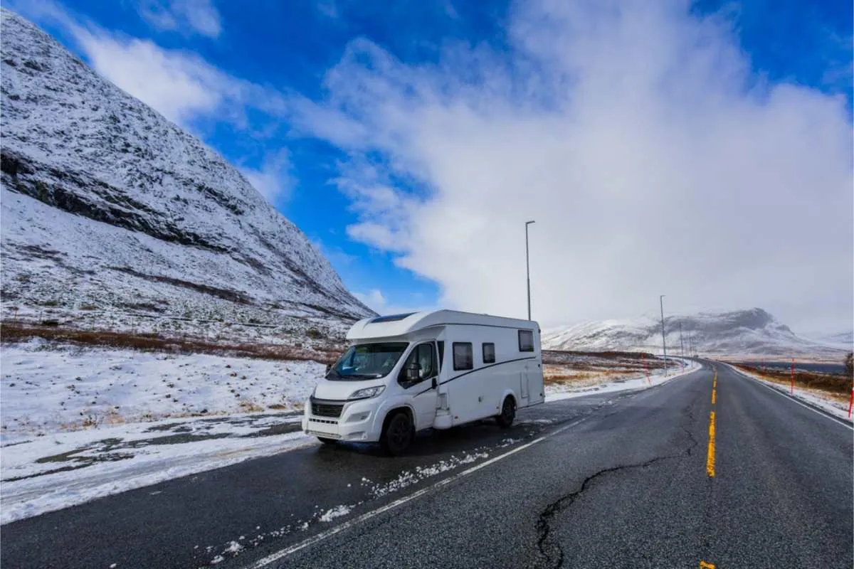 Ein weißes Wohnmobil, geparkt auf einer verschneiten Straße neben einem Berg unter blauem Himmel mit Wolken. Die Straße schlängelt sich in die Ferne und schafft eine beschauliche Szenerie.