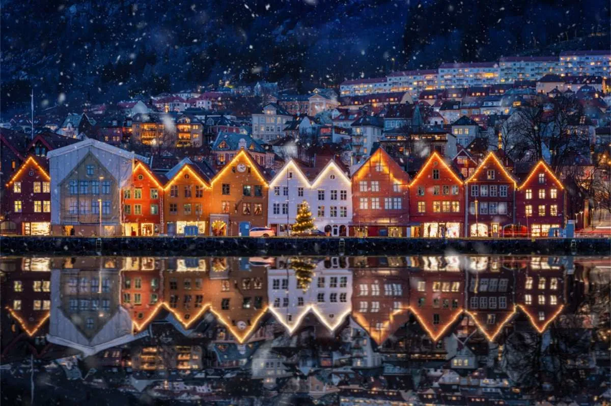 Fargerike hus med festlig belysning speiler seg i et rolig vannspeil, mens snøen faller i Bergen.