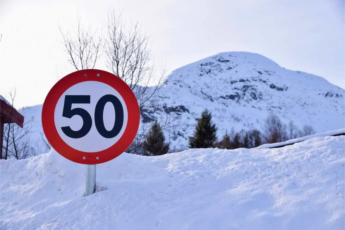 Geschwindigkeitsbegrenzungsschild mit der Angabe „50“ in einer verschneiten Landschaft vor einem Bergpanorama und Bäumen.