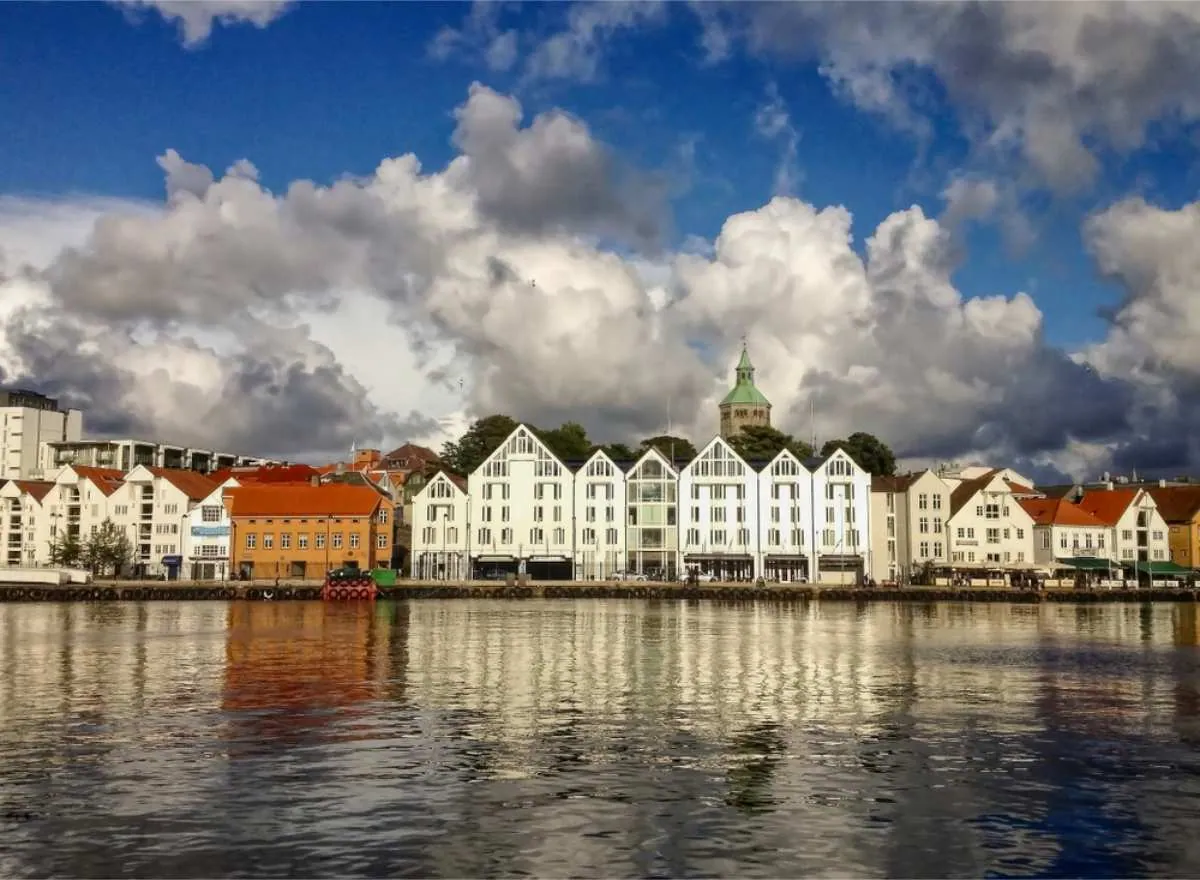 Hvite og oransje bygninger ligger på rekke langs havnefronten i Stavanger under en himmel fylt av dramatiske skyer. Et tårn med grønn topp er synlig.